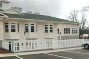 Exterior view of a light beige senior living facility building with multiple windows and a green roof. A white fence runs along the front of the building, and a parking lot with marked spaces is visible in the foreground. Trees without leaves are seen in the background under a cloudy sky.
