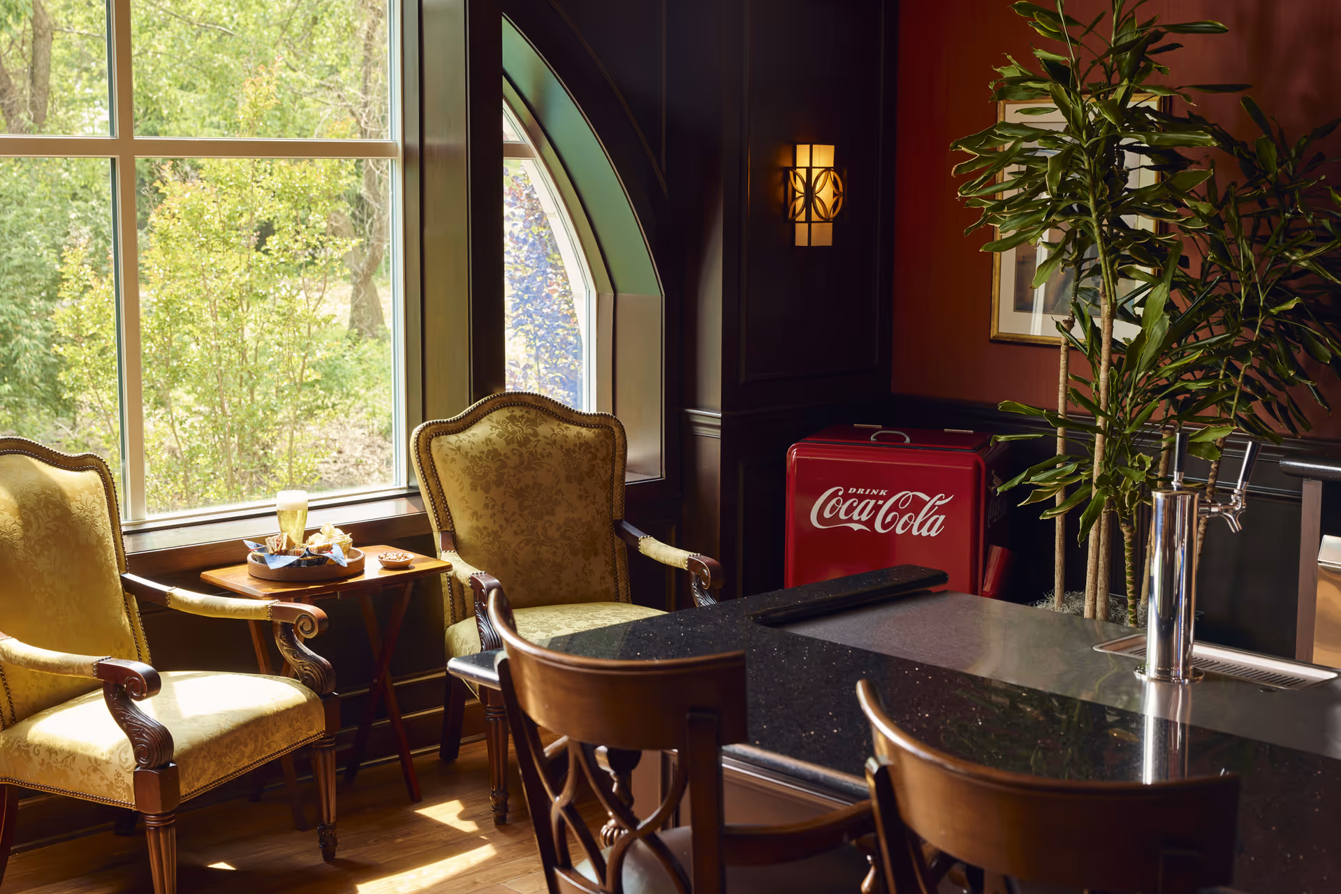A cozy seating area with two ornate upholstered armchairs next to a window overlooking greenery. A small wooden table between the chairs holds a tray with a glass of beer and snacks. In the background, there is a vintage red Coca-Cola cooler and a tall green plant. In the foreground, there is a dark granite countertop with wooden bar stools and a beer tap.