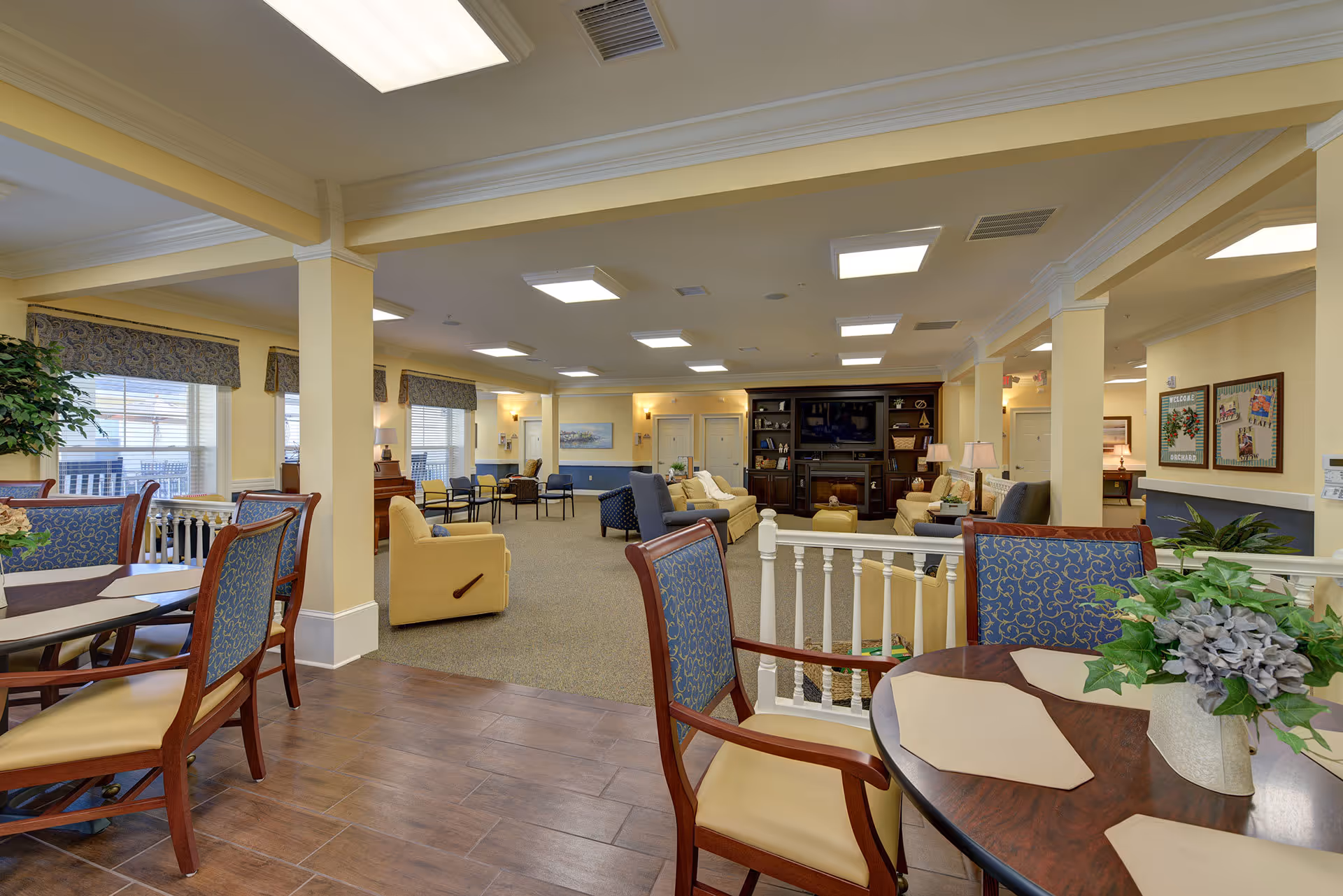 A spacious and well-lit common area in a senior living facility featuring a seating area with sofas and armchairs arranged around a large wooden entertainment center with a TV. In the foreground, there are dining tables with cushioned chairs and placemats, along with a decorative plant on one table. The room has large windows with patterned valances, beige walls, and a mix of carpet and wood flooring.