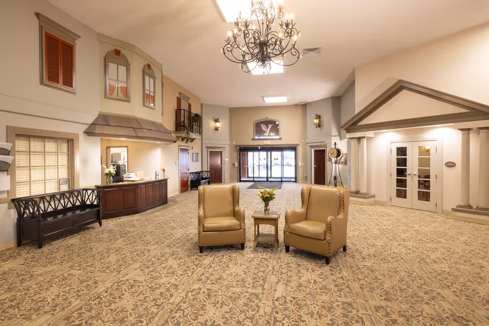 Spacious and well-lit lobby area with two beige armchairs and a small table with flowers in the center. The room features a patterned carpet, a chandelier hanging from the ceiling, decorative wall elements resembling windows and a small balcony, and double glass doors at the far end. There is a reception desk on the left side and a doorway with columns on the right.