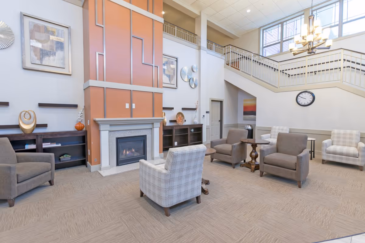 A spacious and well-lit living room area with a high ceiling, featuring a modern fireplace with an orange and gray accent wall. The room is furnished with several armchairs in neutral tones arranged around small wooden tables. There are decorative items on shelves, abstract wall art, a large clock on the wall, and a chandelier hanging from the ceiling. A staircase with a railing leads to an upper level with large windows letting in natural light.