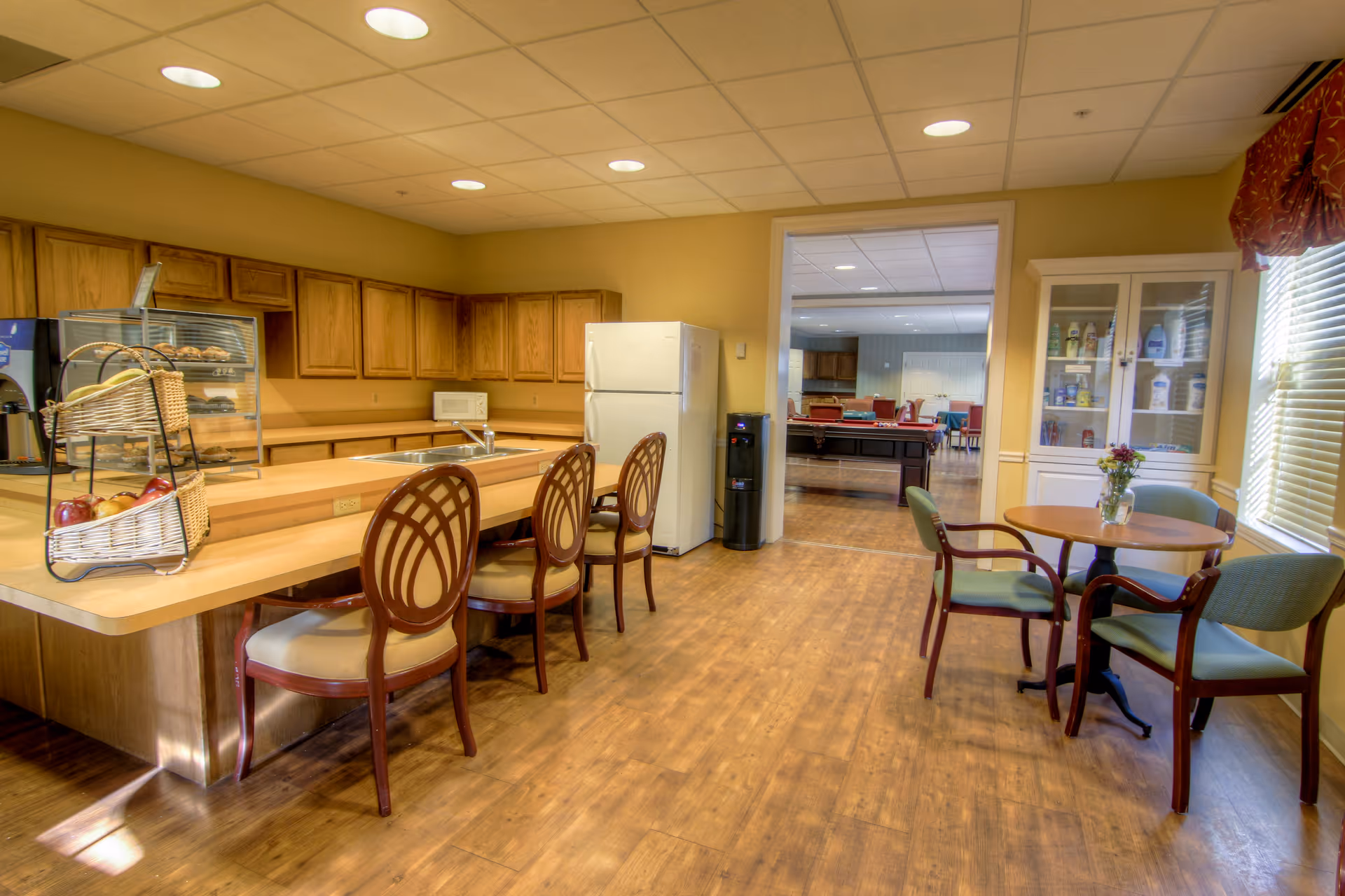 A bright kitchen and dining area in a senior living facility featuring wooden cabinets, a white refrigerator, a microwave, a sink, and a counter with three chairs. A small round table with four chairs and a vase with flowers is near a window with blinds and a red valance. In the background, there is a view into another room with a pool table and additional seating.