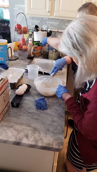 Two elderly individuals wearing gloves are preparing food in a kitchen. One person is whisking a mixture in a bowl on a marble countertop, which also has eggs, a measuring cup, and various kitchen items. The background shows kitchen cabinets, a fruit basket, and other kitchen utensils.
