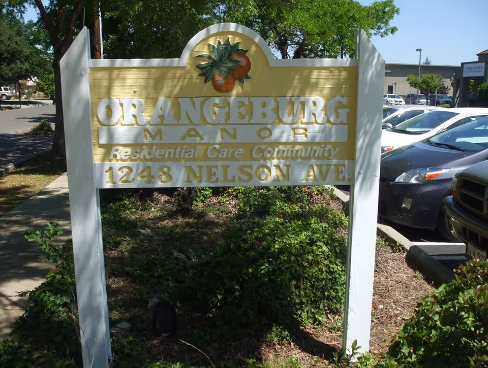 A yellow and white outdoor sign for Orangeburg Manor Residential Care Community located at 1248 Nelson Ave, with a graphic of two oranges and green leaves at the top. The sign is positioned on a grassy area next to a sidewalk and a parking lot with several cars.