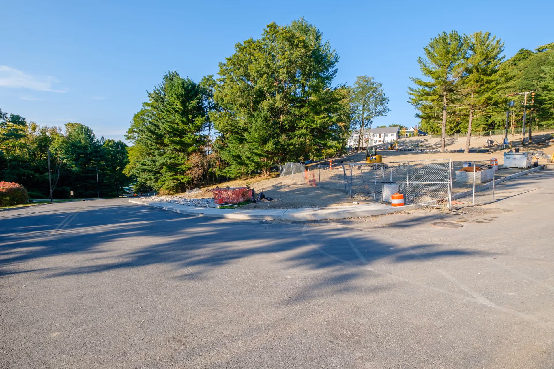 A paved road curves to the left with a construction site on the right side, surrounded by a chain-link fence and orange safety barriers. There are trees and greenery in the background under a clear blue sky.