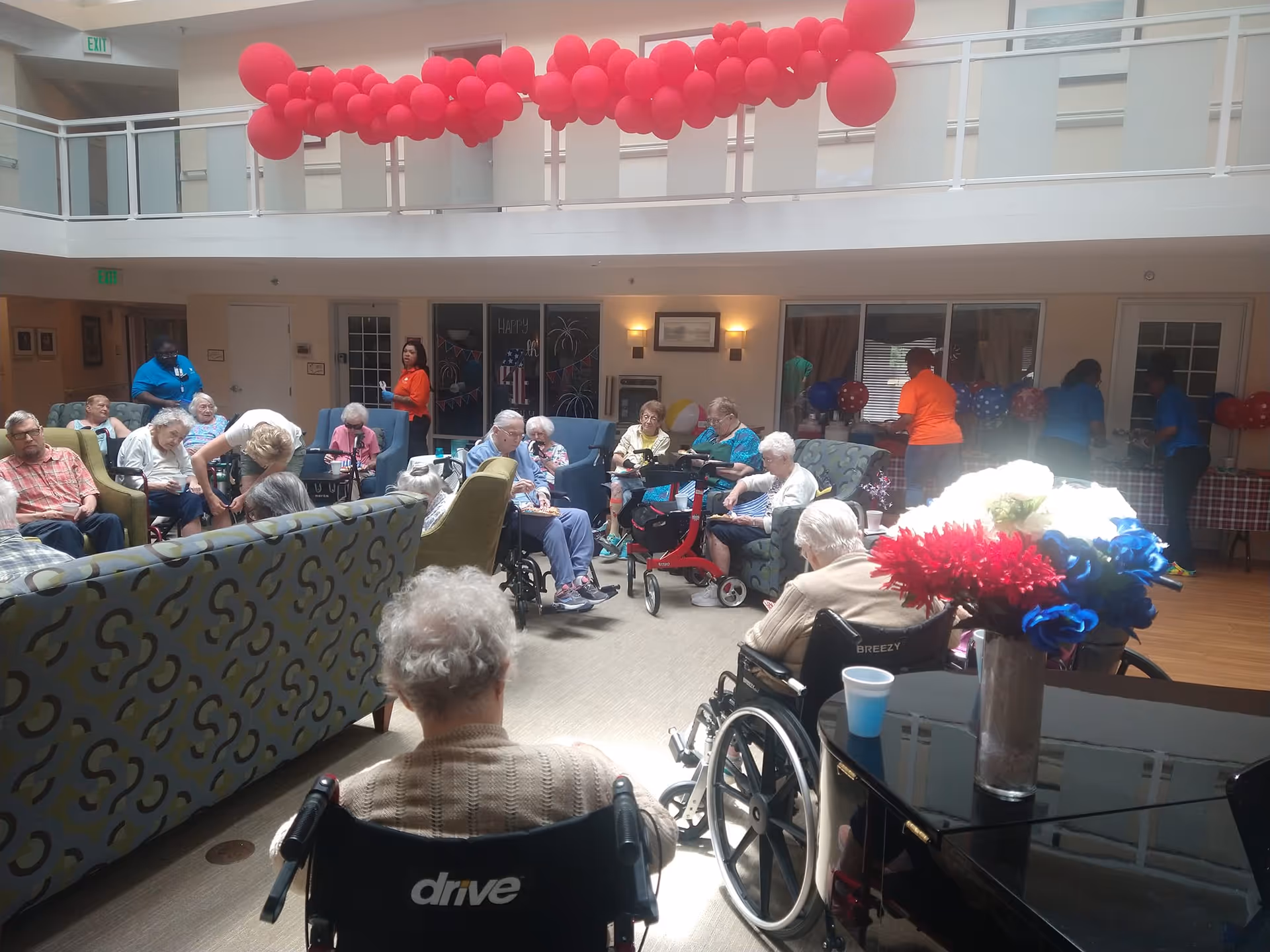 A group of elderly residents sitting in wheelchairs and chairs in a spacious, well-lit common area decorated with red balloons hanging from the upper balcony. Staff members are standing and interacting with the residents, and a table with red, white, and blue decorations is visible in the background. A vase with red, white, and blue flowers is on a piano in the foreground.