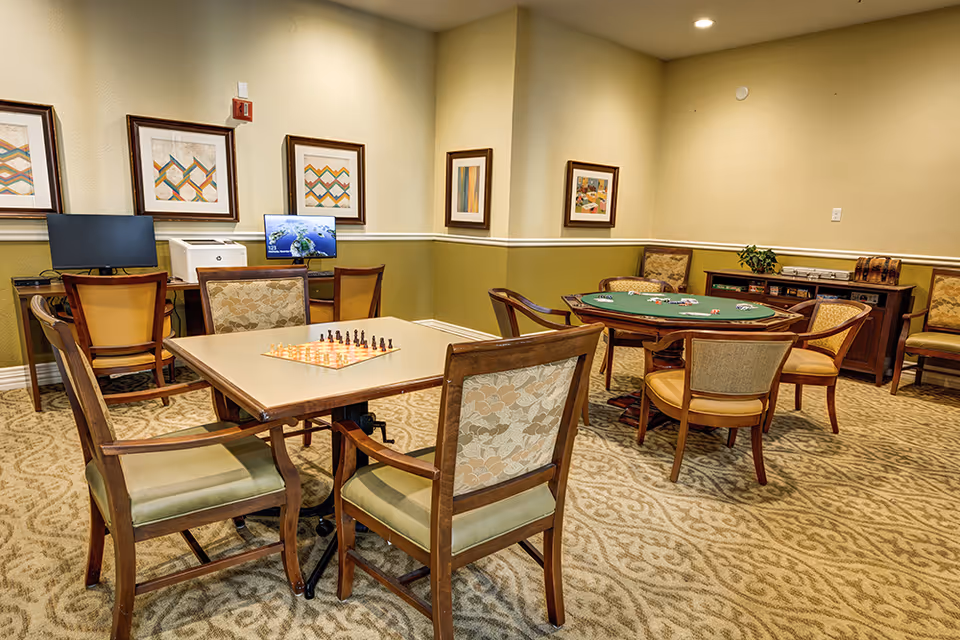 A cozy activity room in a senior living facility featuring two tables with chairs. One table has a chessboard set up, and the other table is a poker table with playing cards and poker chips. The room has patterned carpet, framed artwork on the walls, and a computer workstation with two monitors and a printer in the background.