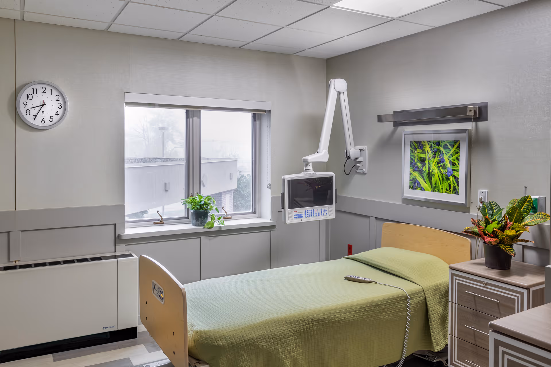 A single hospital-style bed with a green bedspread in a small, clean patient room. The room has a window with a potted plant on the windowsill, a wall-mounted medical monitor, a clock showing 8:35, a framed picture of green plants on the wall, and a bedside table with another potted plant.