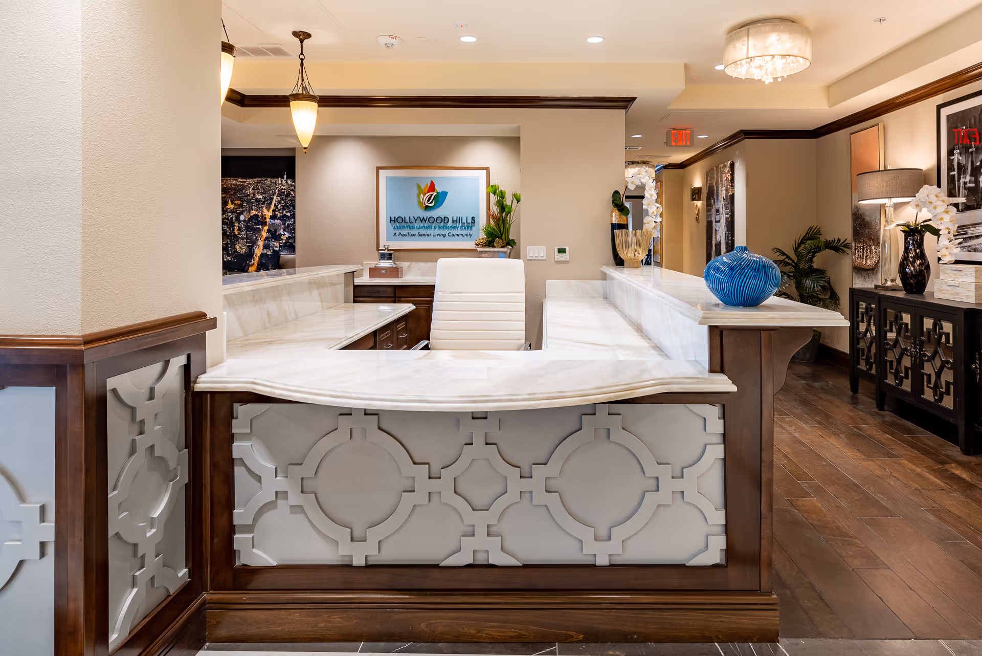 Reception desk area in Hollywood Hills Senior Living facility with a white chair behind the marble counter, decorative vases, plants, and framed artwork on the walls. The space has warm lighting and wooden flooring.