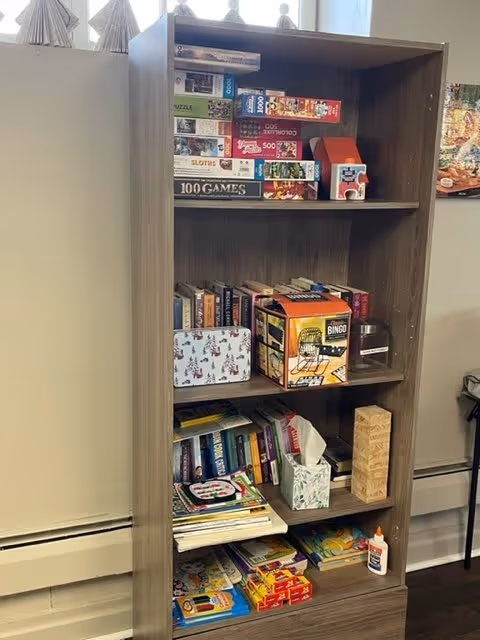 A wooden bookshelf filled with various board games, puzzles, books, a bingo game, a Jenga game, tissues, and craft supplies, located in a room with light-colored walls and a window above the shelf.