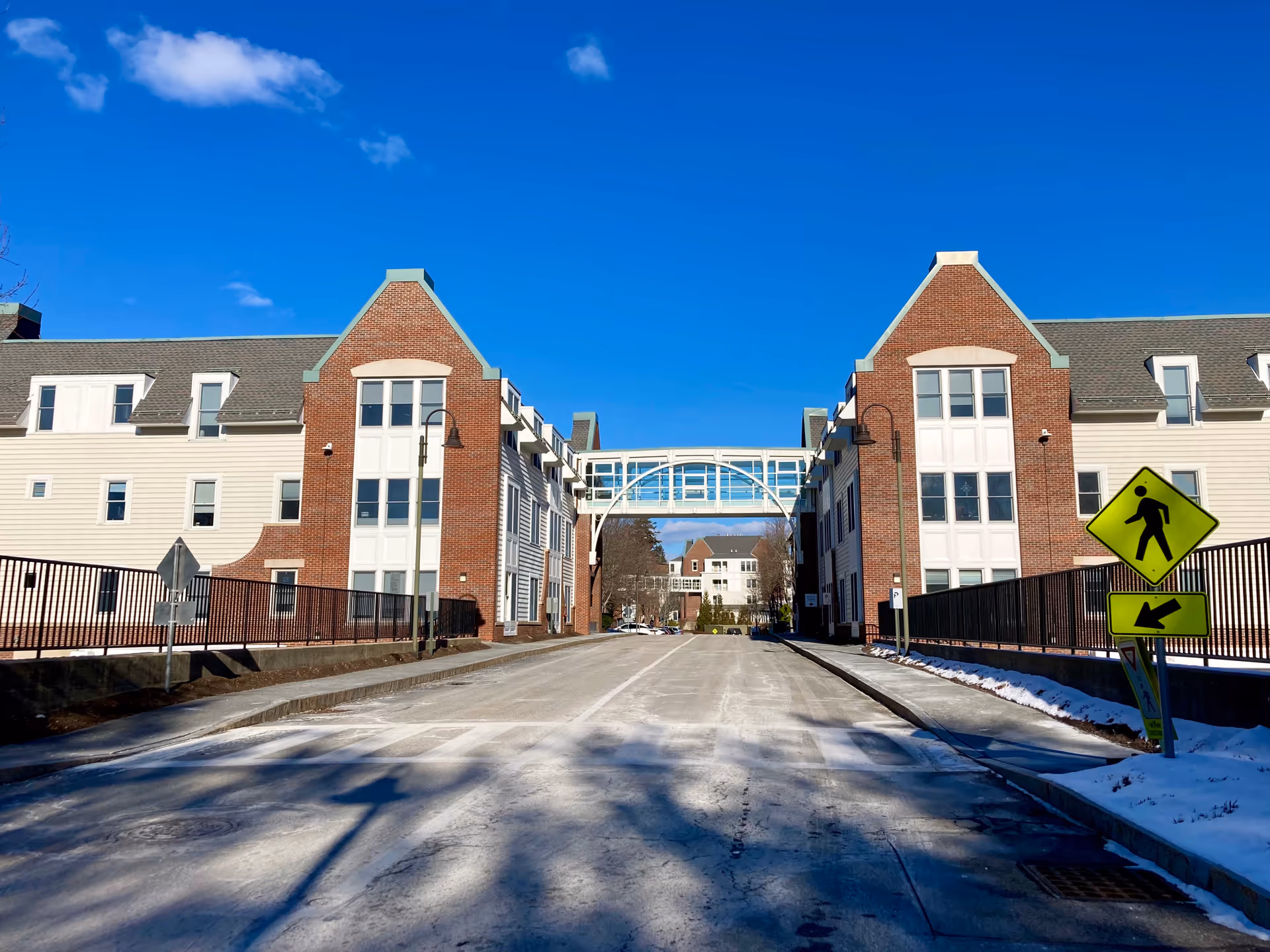 View of a wide driveway or road flanked by two large brick and white-paneled buildings connected by a glass-enclosed pedestrian bridge. There are street lamps and pedestrian crossing signs on the right side. The sky is clear and blue with a few clouds, and there is some snow on the ground edges.