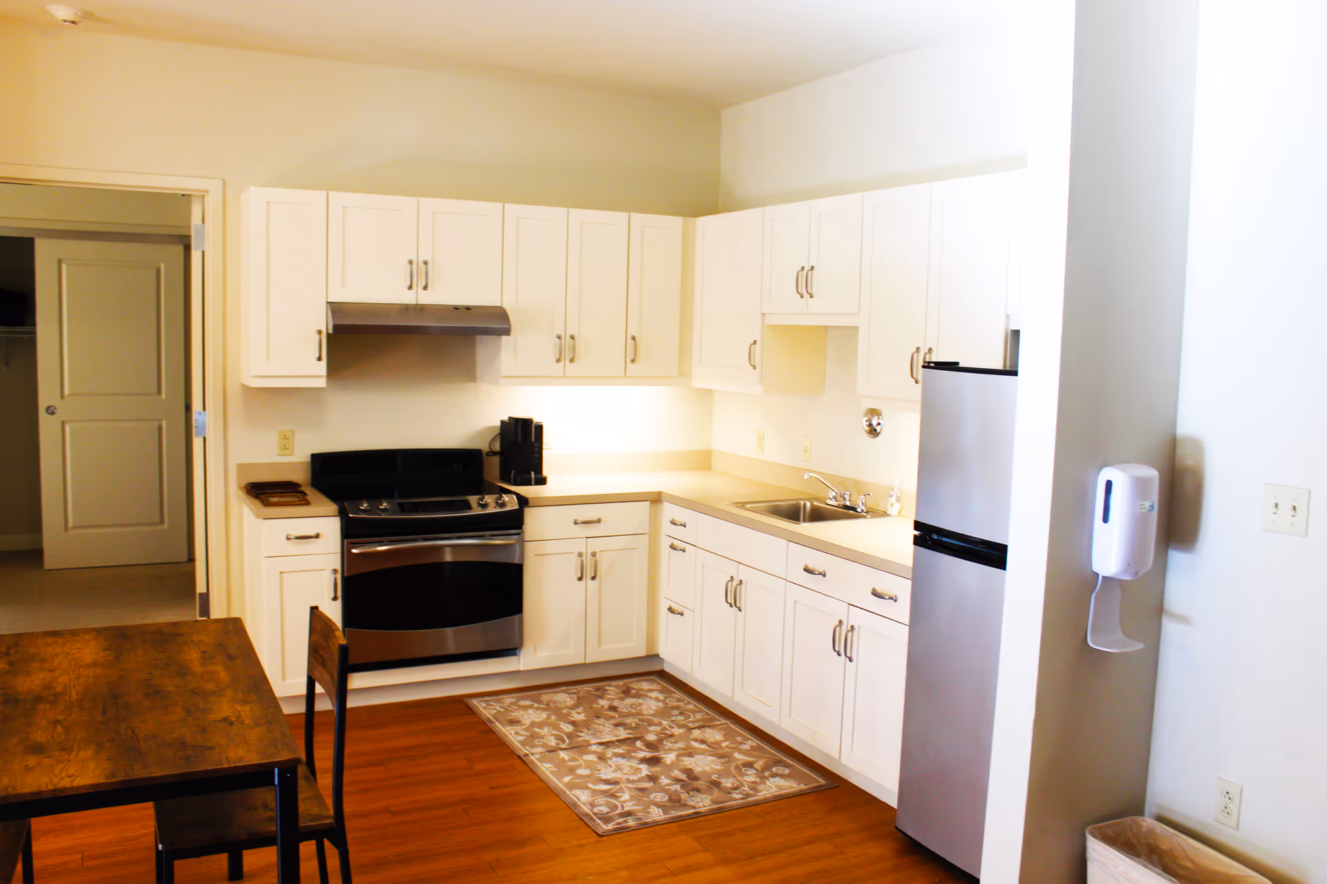 A bright kitchen with white cabinets, a stainless steel stove and refrigerator, a sink, and a wooden dining table with chairs. There is a patterned rug on the wooden floor and a hand sanitizer dispenser mounted on the wall near the refrigerator.