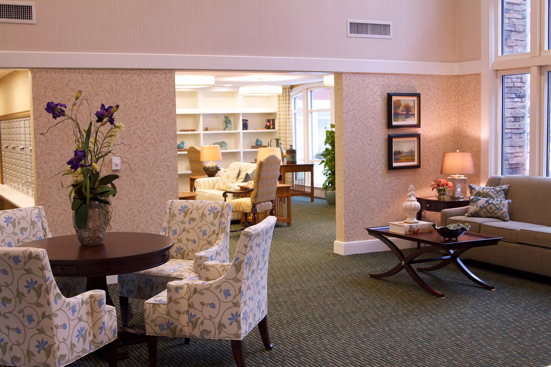 A cozy and well-lit living room area in a senior living facility with floral-patterned chairs around a round wooden table with a vase of purple flowers. In the background, there is a sitting area with armchairs, a sofa, a coffee table, side tables with lamps, framed artwork on the walls, and large windows letting in natural light.
