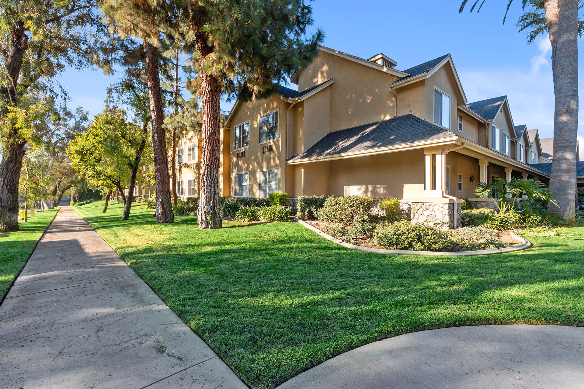 Exterior view of a two-story senior living facility building with beige walls and a dark roof, surrounded by well-maintained green lawns, trees, and shrubs under a clear blue sky.