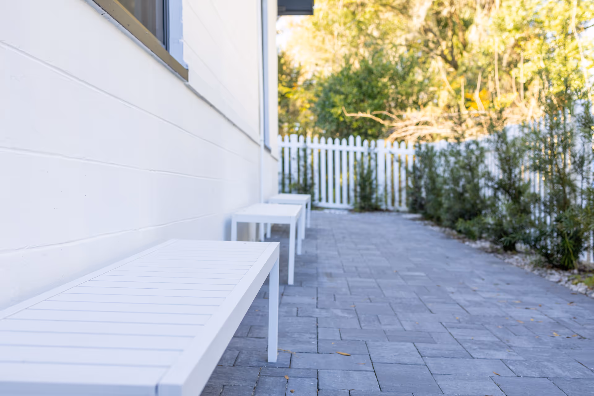 A paved outdoor patio area with white benches lined up against the side of a white building. There is a white picket fence and green shrubs along the edge of the patio, with trees visible in the background.