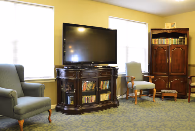 A cozy living room area with two large windows, a flat-screen TV on a dark wooden cabinet filled with books, two upholstered armchairs, and a wooden armoire with books on top.
