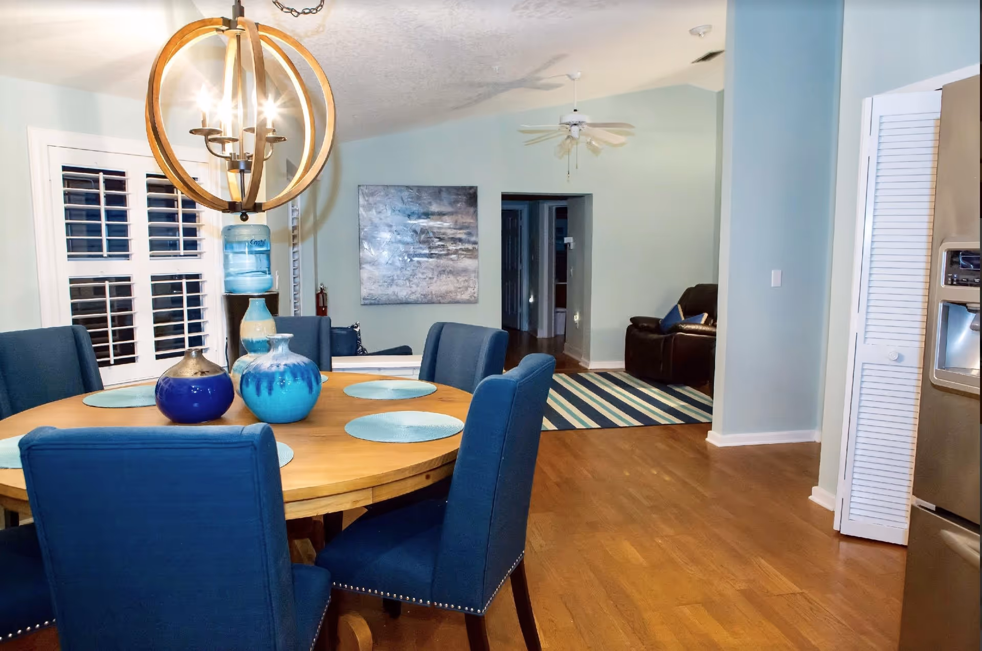 Interior view of a dining area with a round wooden table surrounded by six blue upholstered chairs. The table is set with blue placemats and decorative blue vases. A modern chandelier with exposed bulbs hangs above the table. In the background, there is a living area with a striped rug, a dark leather armchair, a ceiling fan, and a large abstract painting on a light blue wall. Hardwood flooring runs throughout the space.