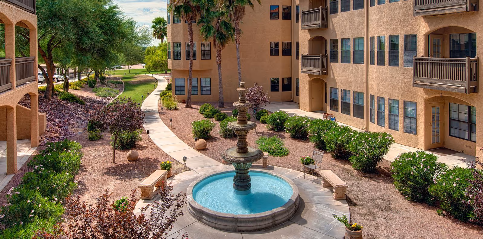 Outdoor courtyard area of a senior living facility with a three-tiered water fountain in the center, surrounded by benches and greenery. The courtyard is bordered by a beige multi-story building with balconies and windows. A winding paved pathway leads through landscaped bushes, trees, and desert-style rocks under a partly cloudy sky.