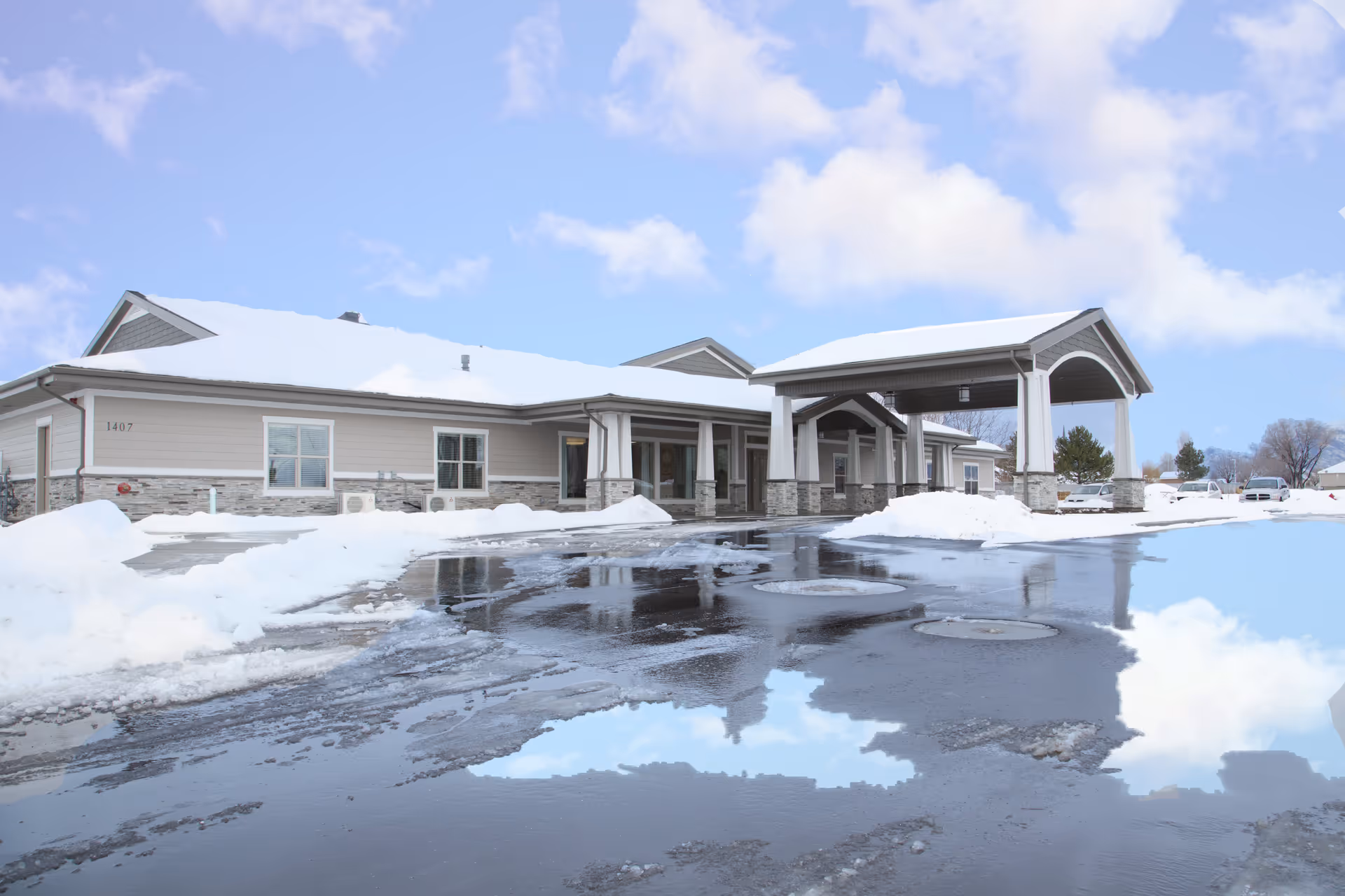 Exterior view of a single-story building with a covered entrance and snow on the ground and roof. The sky is blue with some clouds, and there are a few parked vehicles in the background.