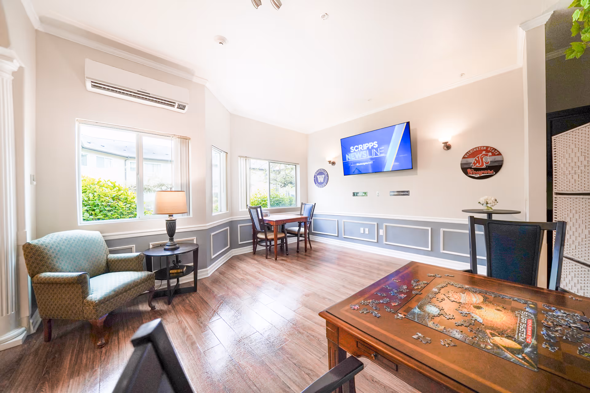 A bright and airy common area in an assisted living facility with large windows letting in natural light. The room features a comfortable armchair next to a small round table with a lamp, a wooden table with chairs, and a puzzle in progress on another wooden table. A flat-screen TV is mounted on the wall displaying 'SCRIPPS NEWSLINE'. The walls are decorated with sports-themed plaques and the floor is wooden.