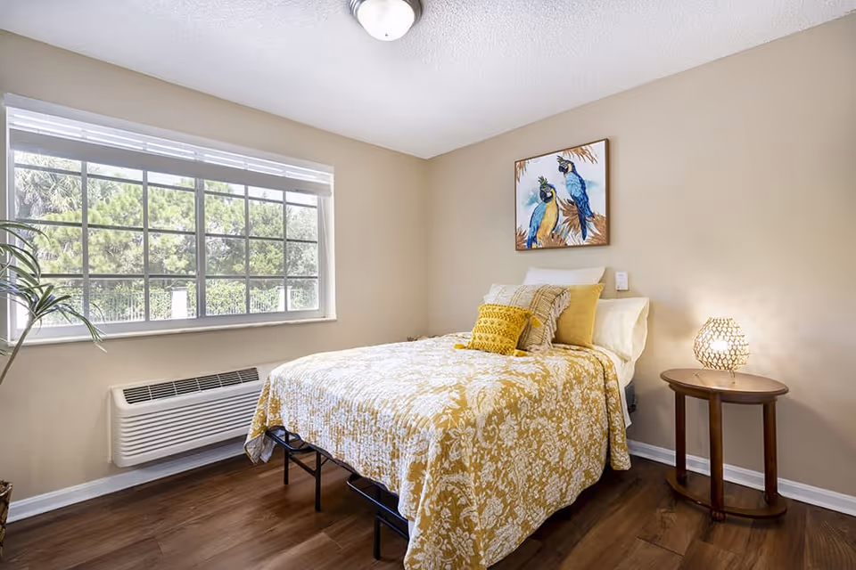 A bright bedroom with a large window showing greenery outside. The room features a bed with a yellow and white floral bedspread, several pillows, a small round wooden side table with a decorative lamp, and a framed picture of two colorful birds on the wall above the bed. The floor is wooden, and the walls are painted beige.