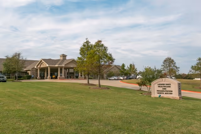 Exterior view of Legend Oaks Healthcare and Rehabilitation - North Austin facility showing a large grassy area with a few trees, a driveway leading to the building entrance with a covered drop-off area, and a sign displaying the facility name.