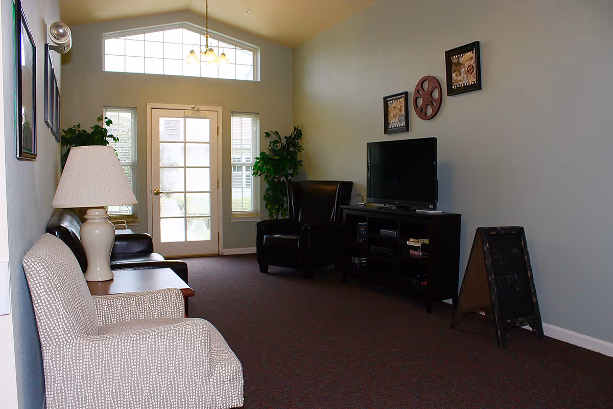 A small living room area with a patterned armchair and a table lamp on the left, a black leather armchair and sofa along the left wall, a black TV stand with a flat-screen TV and some books on the right, and a door with windows letting in natural light at the far end. There are two plants near the door and framed pictures on the walls.