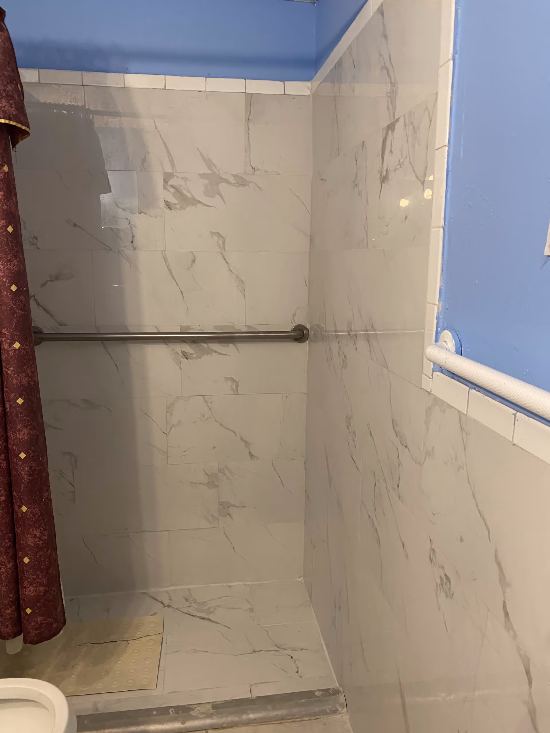 A small shower area with white marble-patterned tiles on the walls and floor, a metal grab bar on the back wall, a maroon shower curtain with a diamond pattern on the left, and a blue painted wall with a white grab bar on the right.