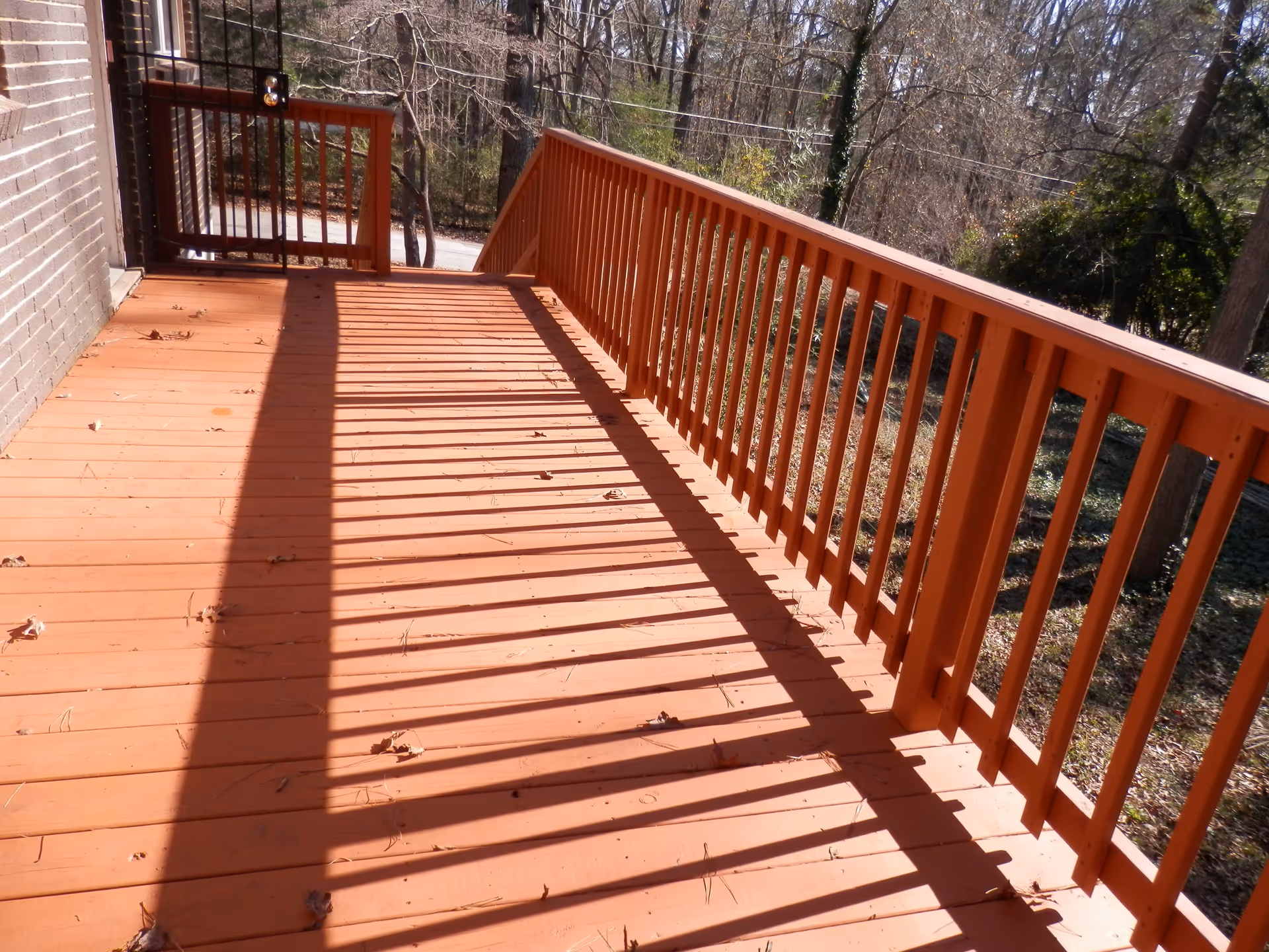 Orange-painted wooden deck with railings casting striped shadows, attached to a brick building and overlooking trees.