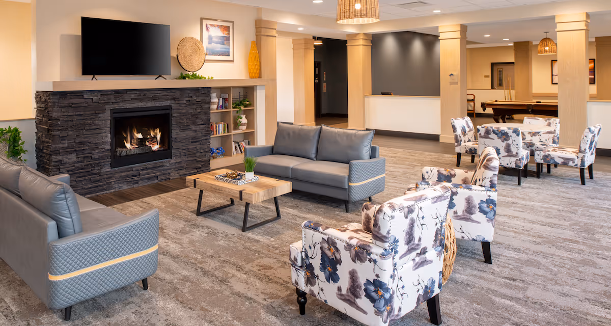 Spacious senior living common area with sofas, floral armchairs, a stone fireplace and TV, coffee table, and a pool table in the background.