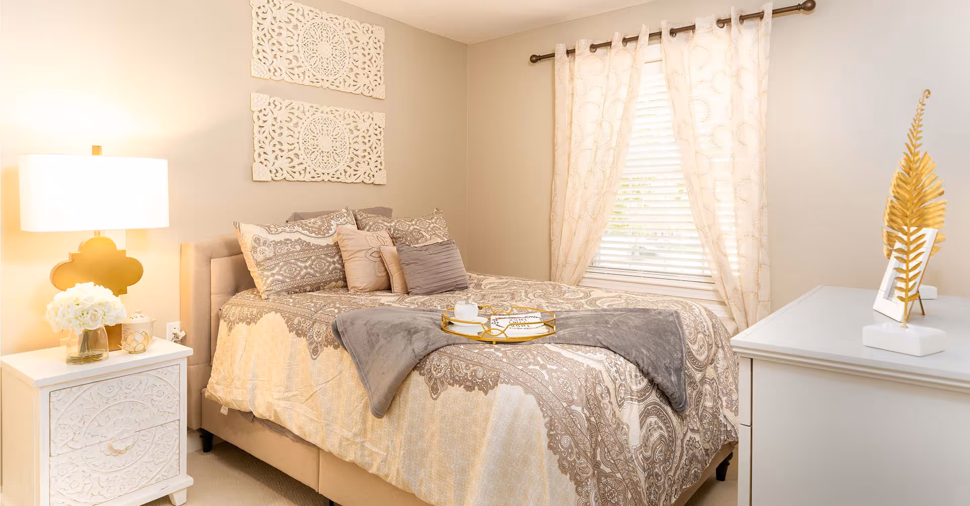 A cozy bedroom with a beige and patterned bedspread, several decorative pillows, and a gray throw blanket. There is a white nightstand with a lamp and a vase of white flowers on the left side of the bed. The wall above the bed features two ornate white wall hangings. A window with light-colored curtains is on the right side, and a white dresser with a gold decorative piece and a photo frame is visible in the foreground.