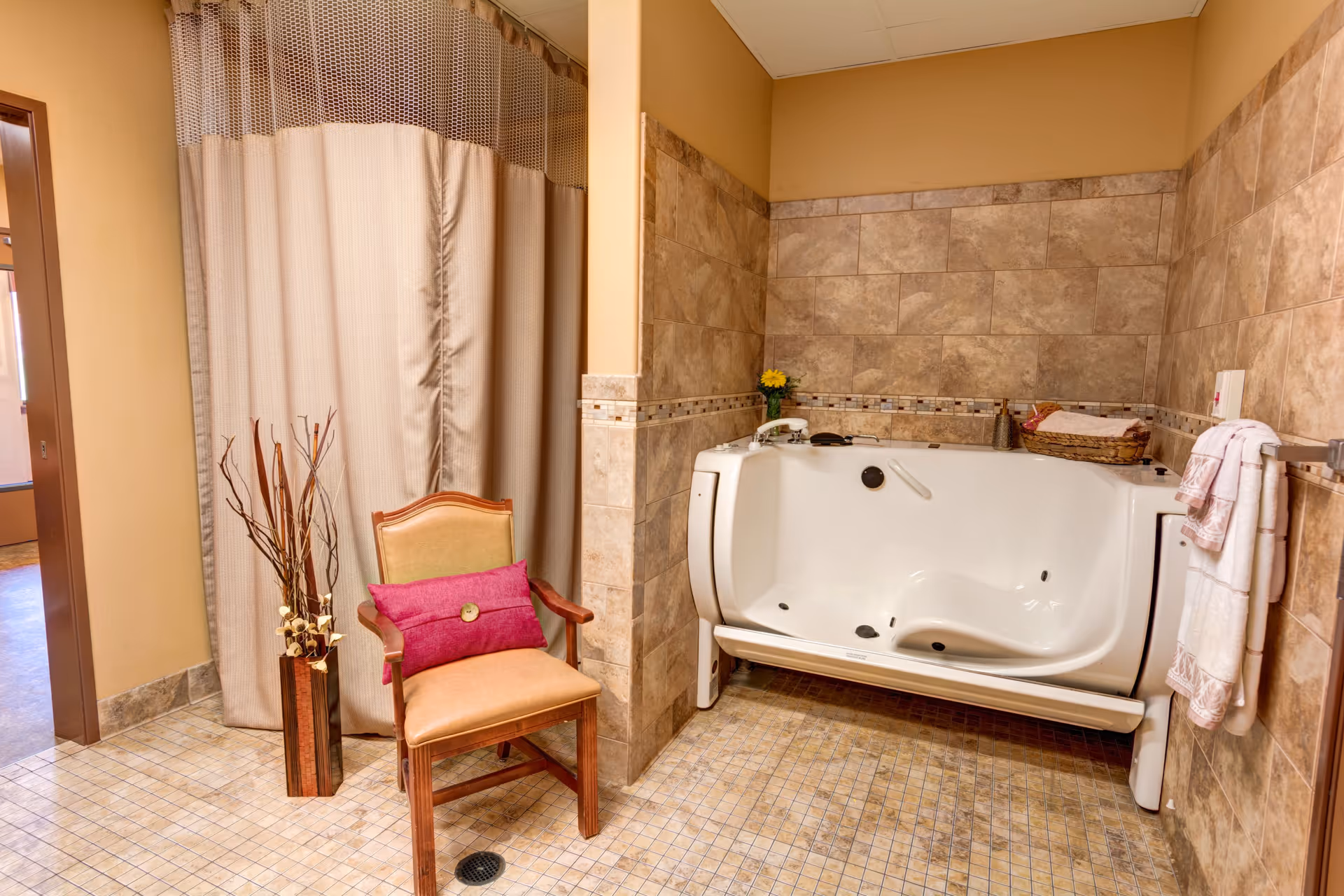 Tiled bathroom featuring a walk-in bathtub, a wooden chair with a pink cushion, and a beige privacy curtain.