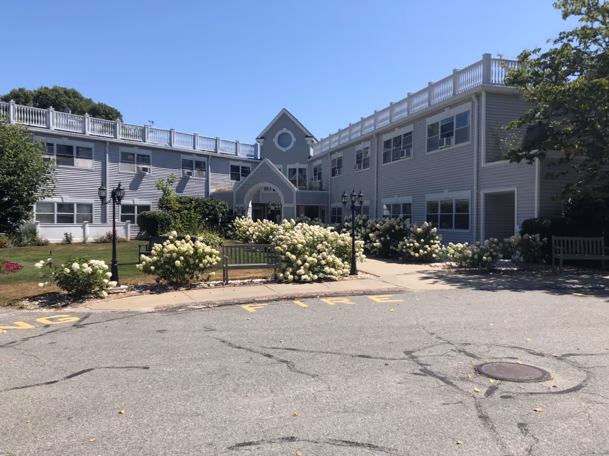 Exterior view of a two-story nursing and rehabilitation center building with gray siding, white trim, and multiple windows. The entrance features an archway and is surrounded by well-maintained landscaping including white flowering bushes, green shrubs, and benches. The sky is clear and blue.