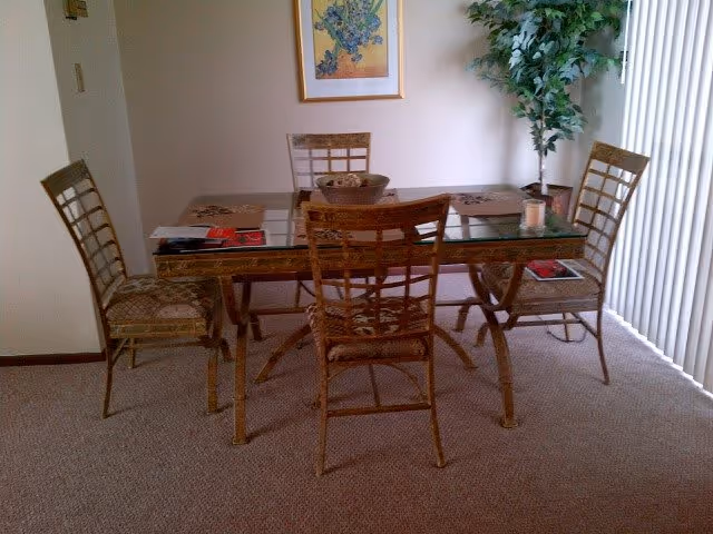 A dining area with a glass-top table and four wooden chairs with lattice backs. The table has placemats, a bowl with decorative items, and a candle. There is a framed picture on the wall and a tall green plant in the corner near vertical blinds covering a window or door.