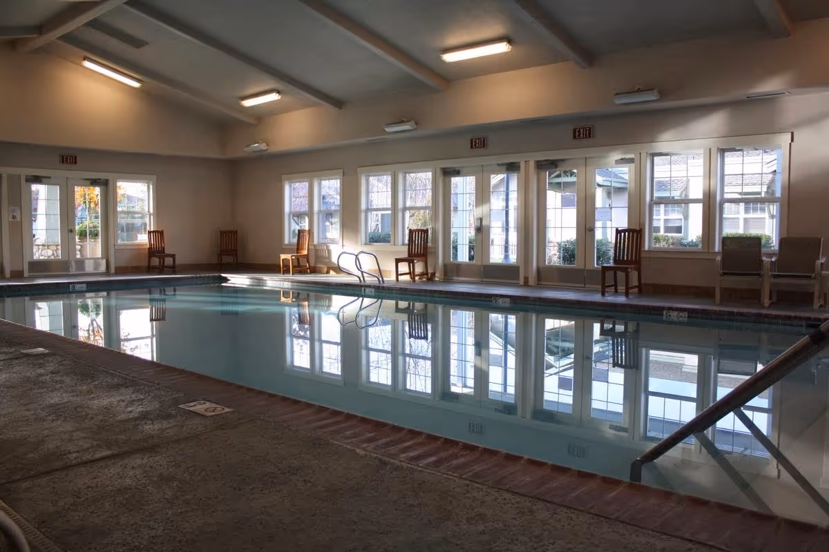Indoor swimming pool with clear water reflecting the windows and ceiling lights. Several wooden chairs and lounge chairs are placed along the walls near large windows and glass doors. The room has a high ceiling with multiple light fixtures and exit signs above the doors.