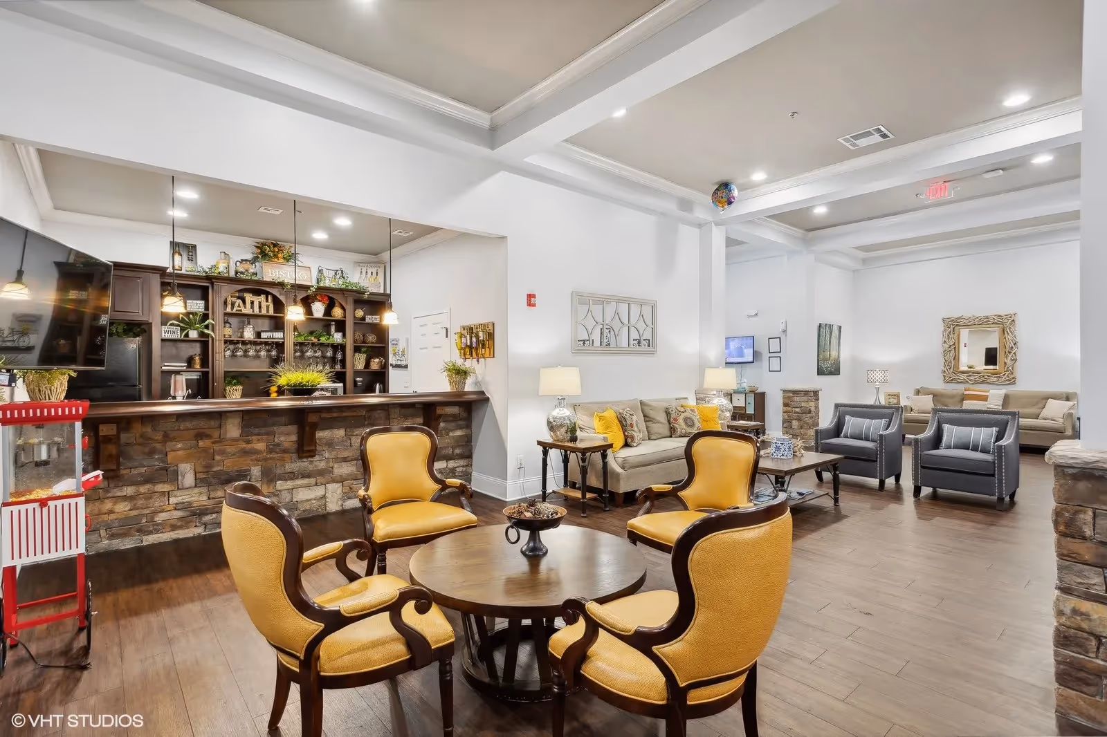 A spacious and well-lit common area in a senior living facility featuring a seating arrangement with four yellow upholstered chairs around a round wooden table in the foreground. Behind it is a stone-faced counter with shelves displaying decorative items and plants. The room has wooden flooring, white walls, and additional seating areas with sofas and armchairs. The ceiling has recessed lighting and a tray design.
