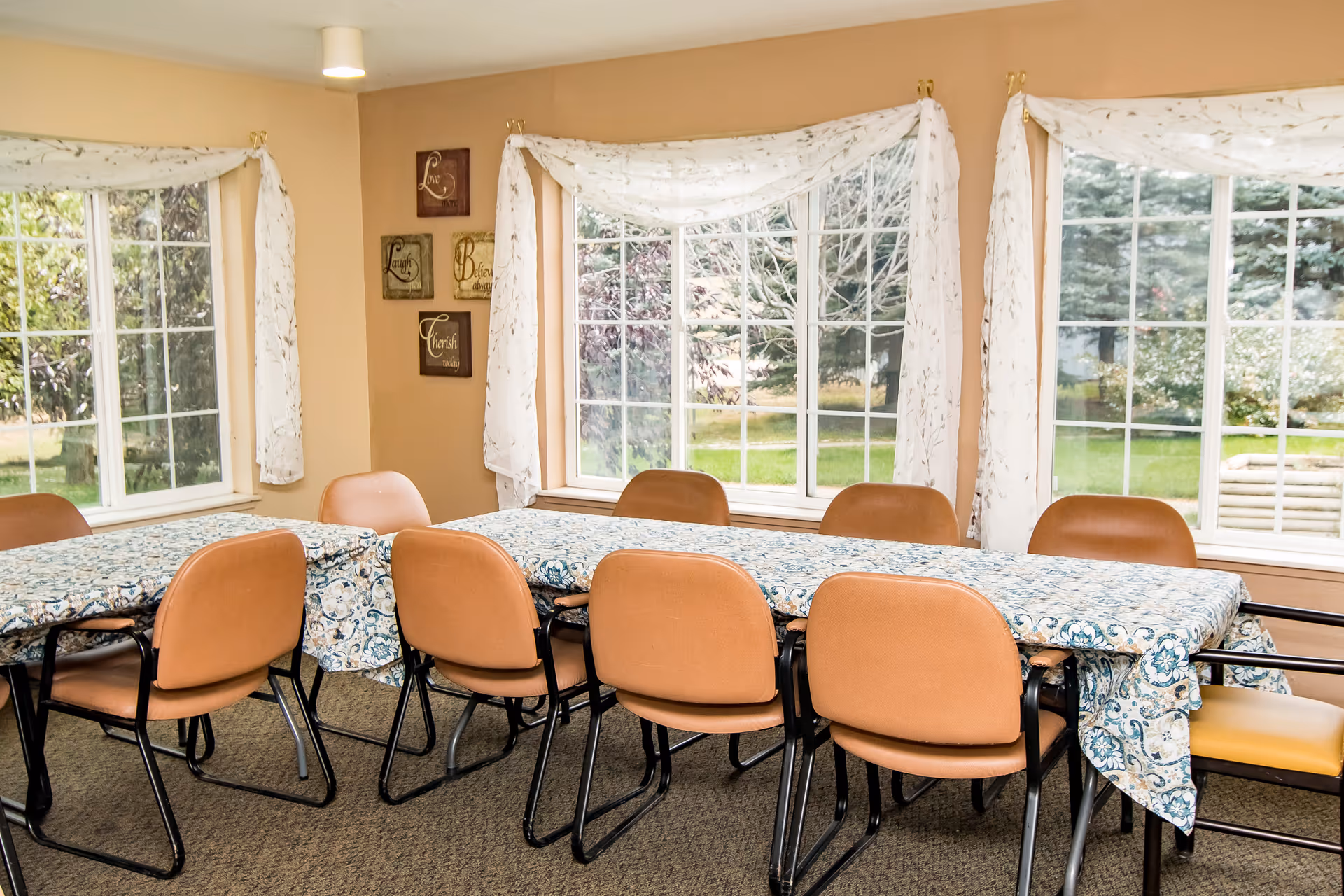 A dining room with two tables covered with patterned tablecloths and surrounded by brown chairs. Large windows with white curtains allow natural light to brighten the room, and outside greenery is visible. Wall decorations with inspirational words are hung on a beige wall.
