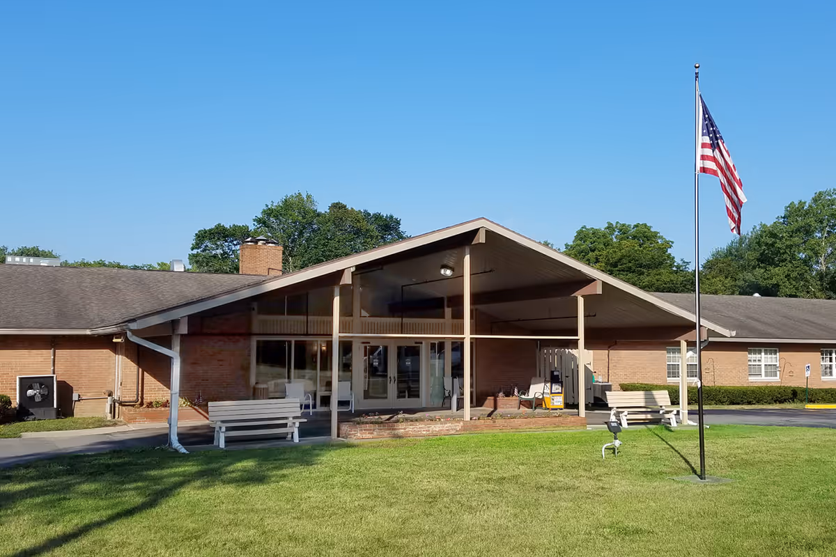 Front exterior view of The Grand Rehabilitation and Nursing at Batavia building with a covered entrance, benches, an American flag on a flagpole, and green lawn in front under a clear blue sky.