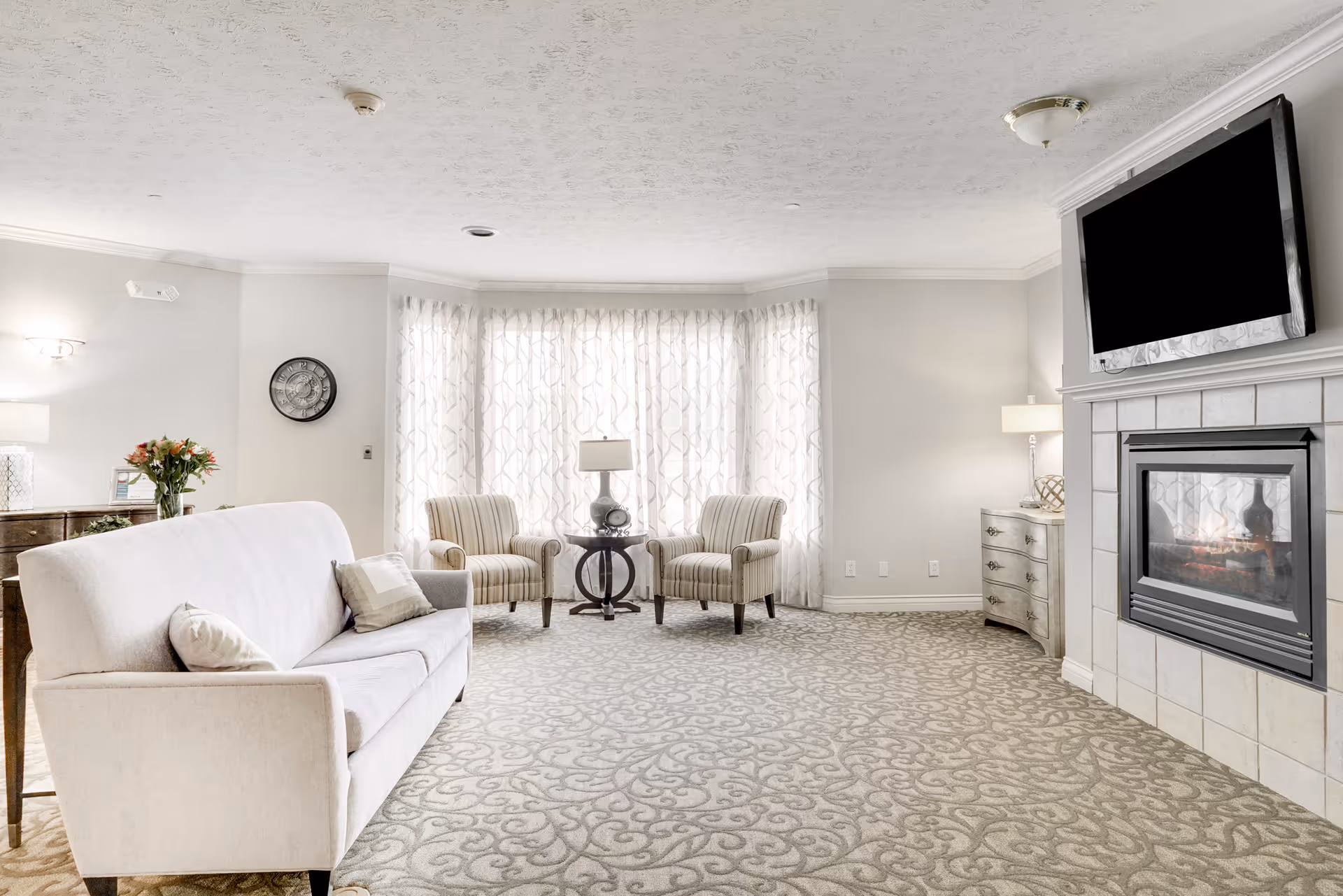 Bright living room with a sofa, two armchairs by a large curtained bay window, patterned carpet, a fireplace and wall-mounted TV.