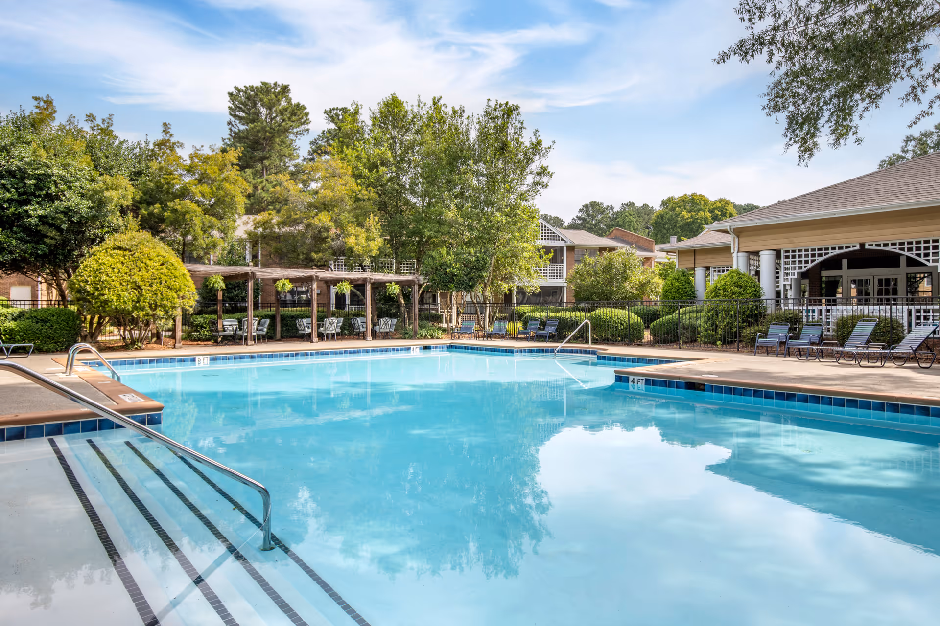Outdoor swimming pool with clear blue water surrounded by lounge chairs, trees, and a pergola with seating. Residential buildings and a covered patio area are visible in the background under a partly cloudy sky.