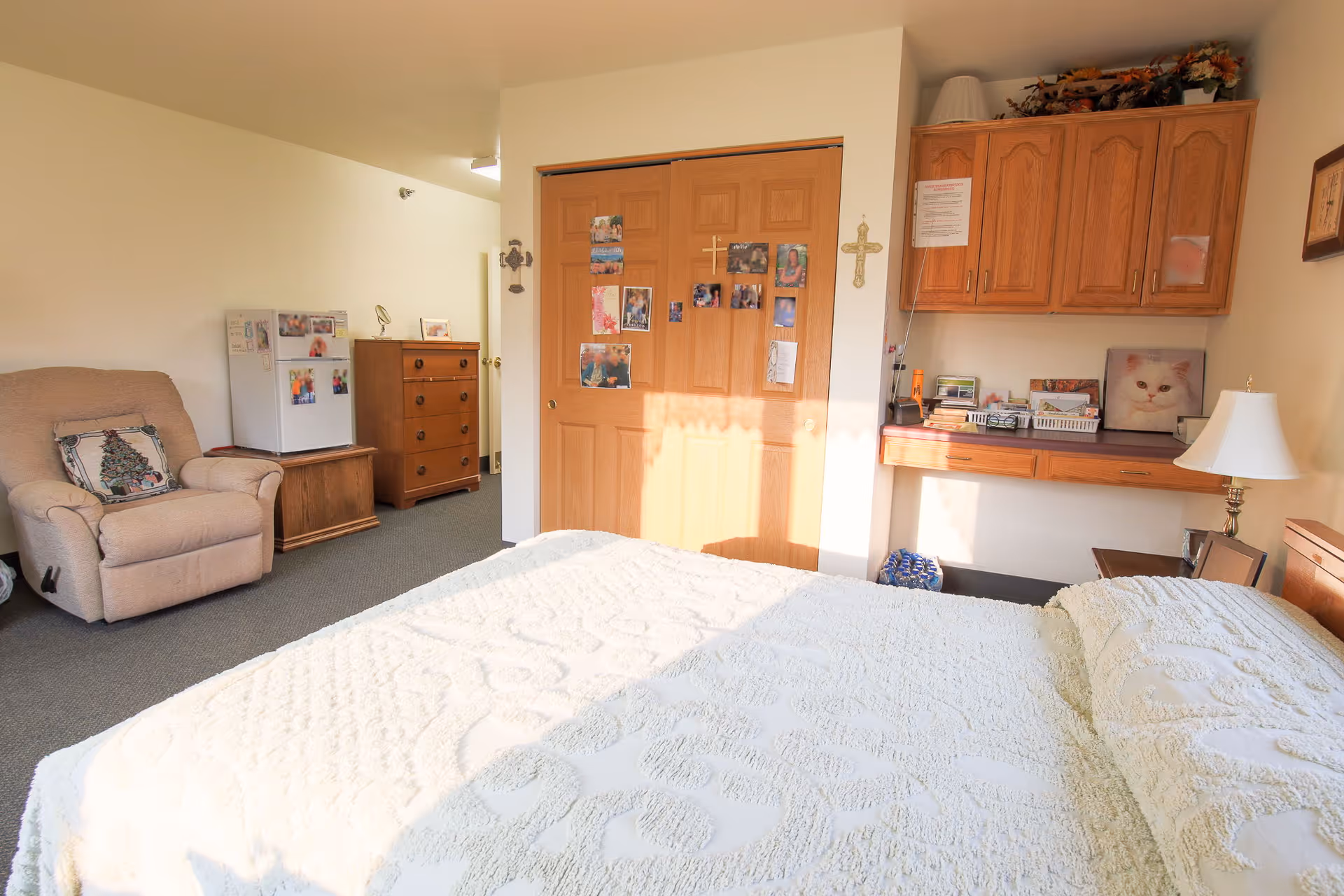 Sunlit bedroom with a bed in the foreground, a recliner, small refrigerator and dresser, closet doors decorated with photos, and built-in cabinets and a desk.