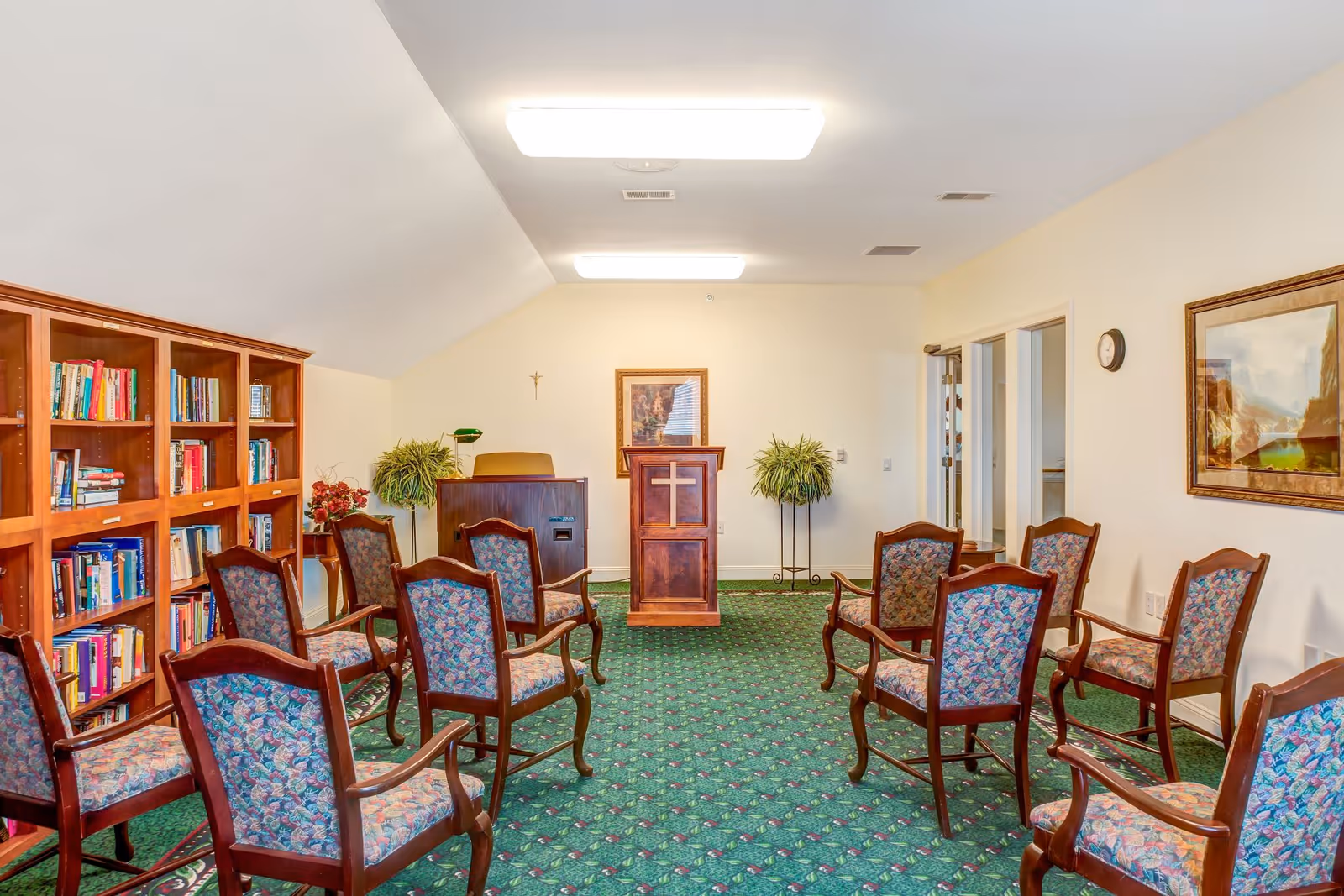 A small chapel or prayer room with a wooden podium featuring a cross, surrounded by several upholstered chairs arranged in rows. There is a wooden bookshelf filled with books on the left side, two potted plants near the podium, a framed picture on the wall, and a clock on the right wall. The room has a green patterned carpet and bright overhead lighting.