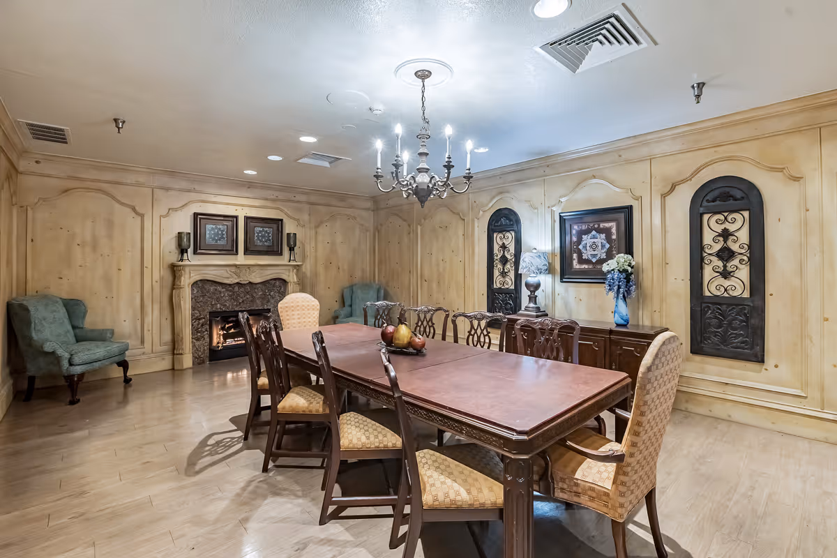A formal dining room with a long wooden table surrounded by eight chairs, two upholstered armchairs at the ends and six wooden chairs with cushioned seats. The room features wood-paneled walls, a decorative fireplace with a fire burning, two green upholstered armchairs in the corners, framed artwork, a chandelier hanging from the ceiling, and decorative wall panels with wrought iron accents.