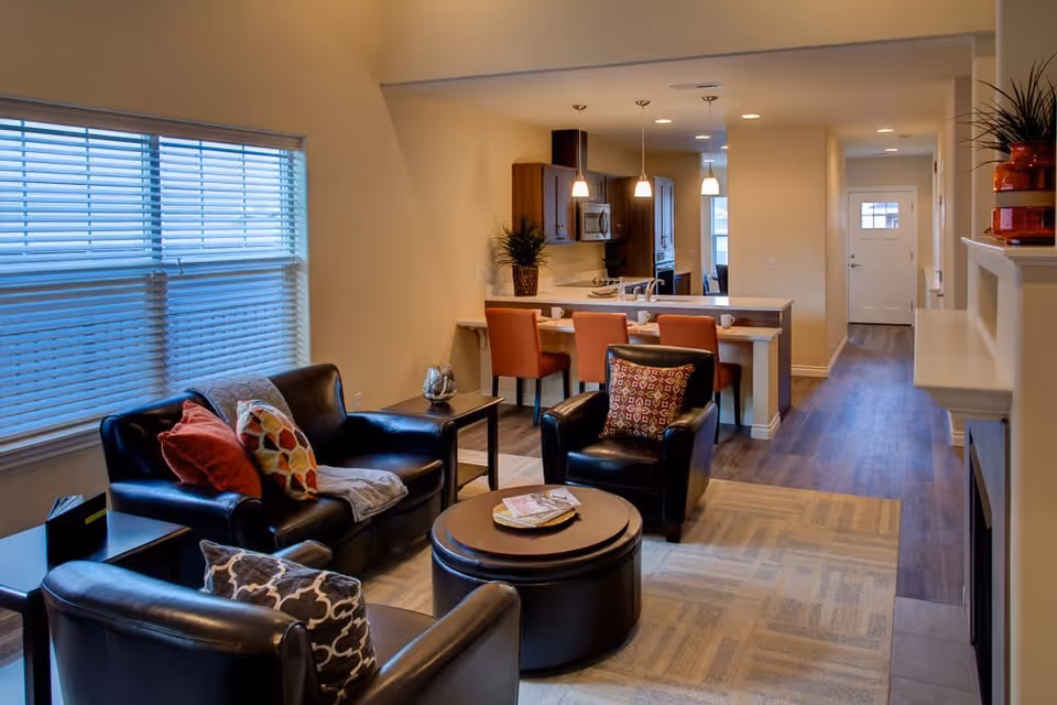 A cozy living room area with three black leather chairs and a matching loveseat, each adorned with colorful throw pillows. A round coffee table with magazines sits in the center on a patterned rug. Behind the seating area is a kitchen with a breakfast bar featuring four orange bar stools. The kitchen has wooden cabinets, a microwave, and pendant lights hanging above the counter. A hallway leads to a white door at the back. The room has warm lighting and a large window with blinds on the left side.