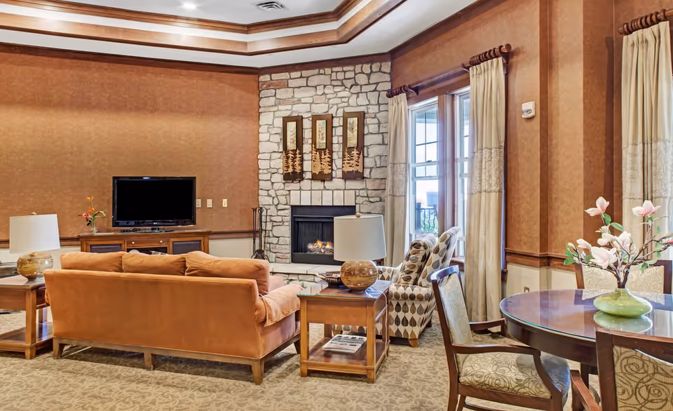 A cozy living room with a brown sofa, patterned armchairs, a wooden coffee table with a lamp, and a flat-screen TV on a wooden stand. The room features a stone fireplace with three decorative wall hangings above it, large windows with beige curtains, and a round wooden dining table with chairs and a vase of flowers.