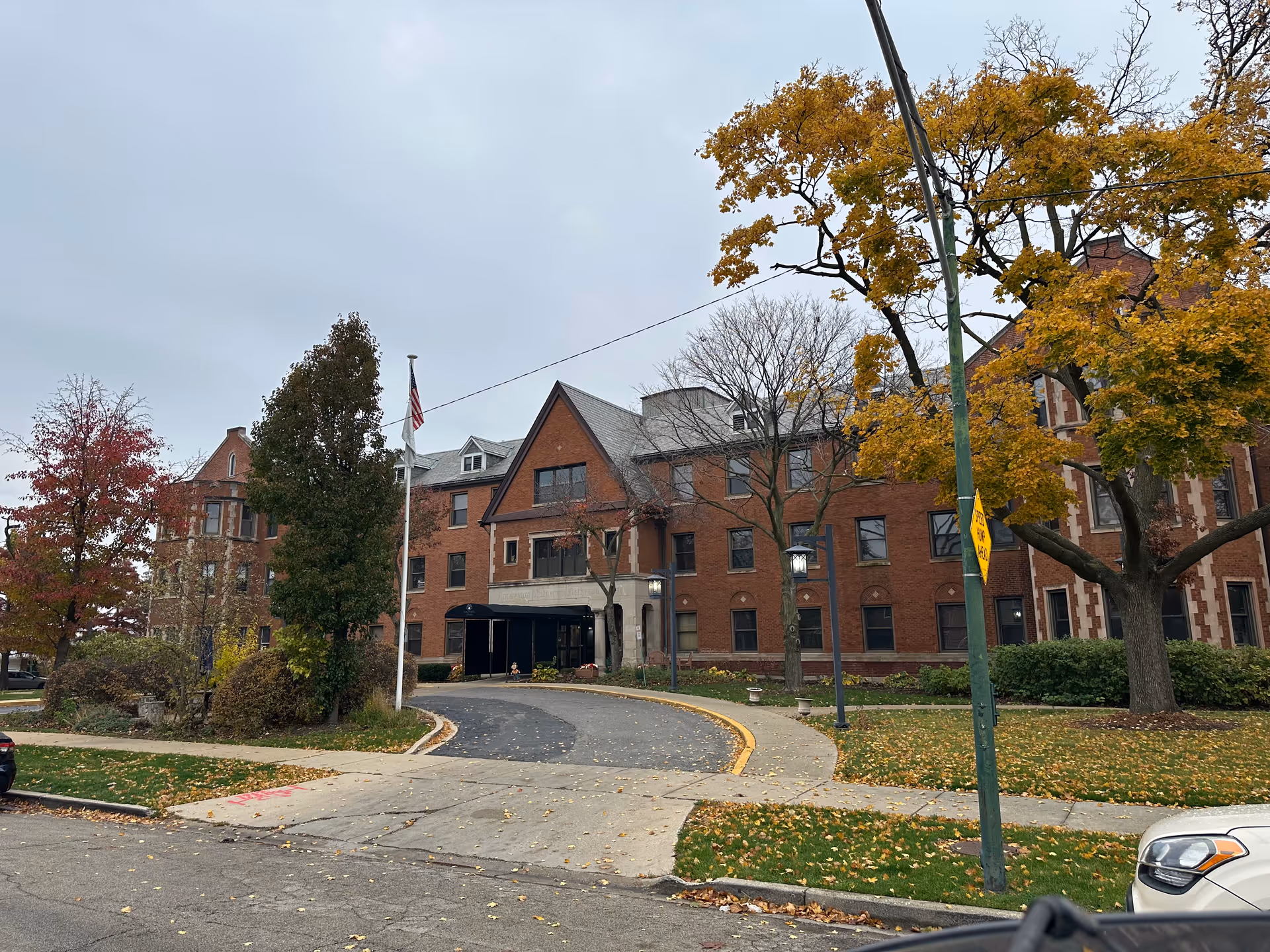 Exterior view of a large brick building with multiple windows and a peaked roof, surrounded by trees with autumn foliage. A curved driveway leads to the entrance, and an American flag is flying on a flagpole near the front. The sky is overcast.
