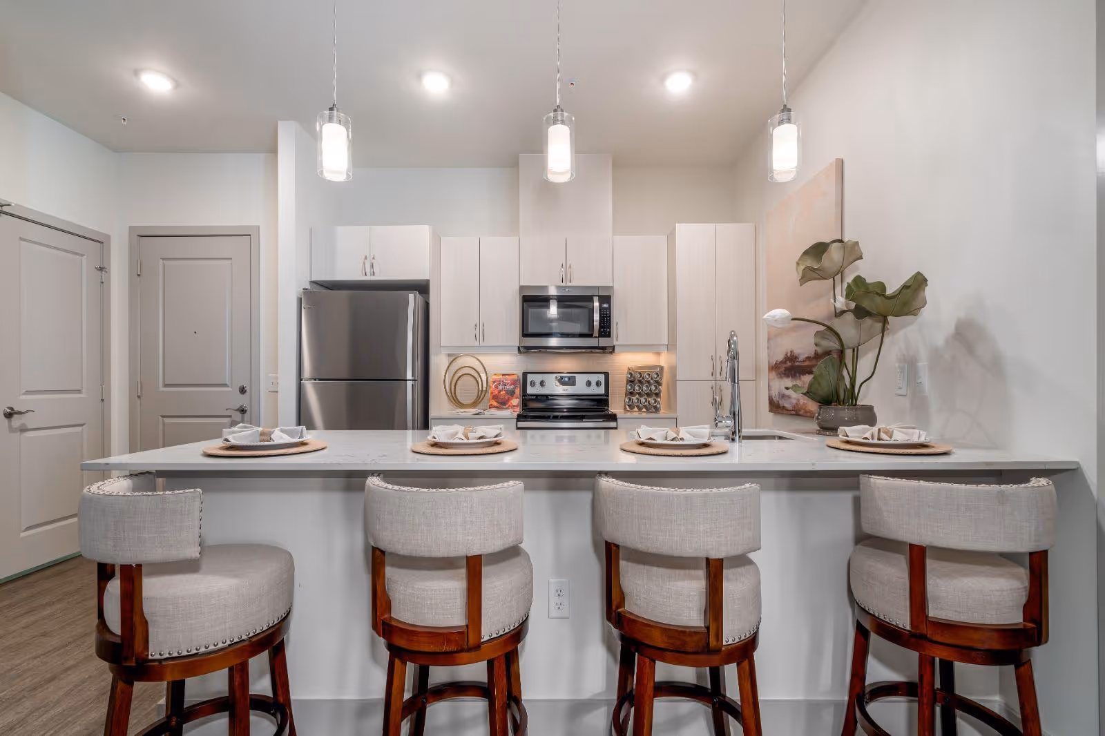 Modern kitchen with a marble island and four upholstered bar stools, stainless steel appliances, pendant lights, and a decorative plant.