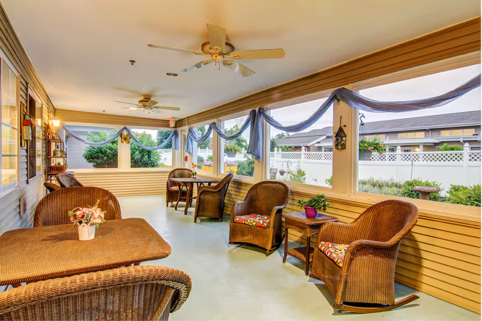 Sunlit enclosed sunroom with wicker chairs and tables, ceiling fans, and large windows overlooking a fenced garden.