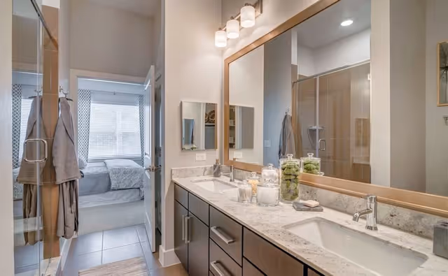 Modern bathroom with a double sink vanity featuring a large mirror and light fixtures above. There are decorative jars and soap on the countertop. A glass shower enclosure is visible on the left, and through an open door, a bedroom with a bed and window with blinds can be seen.