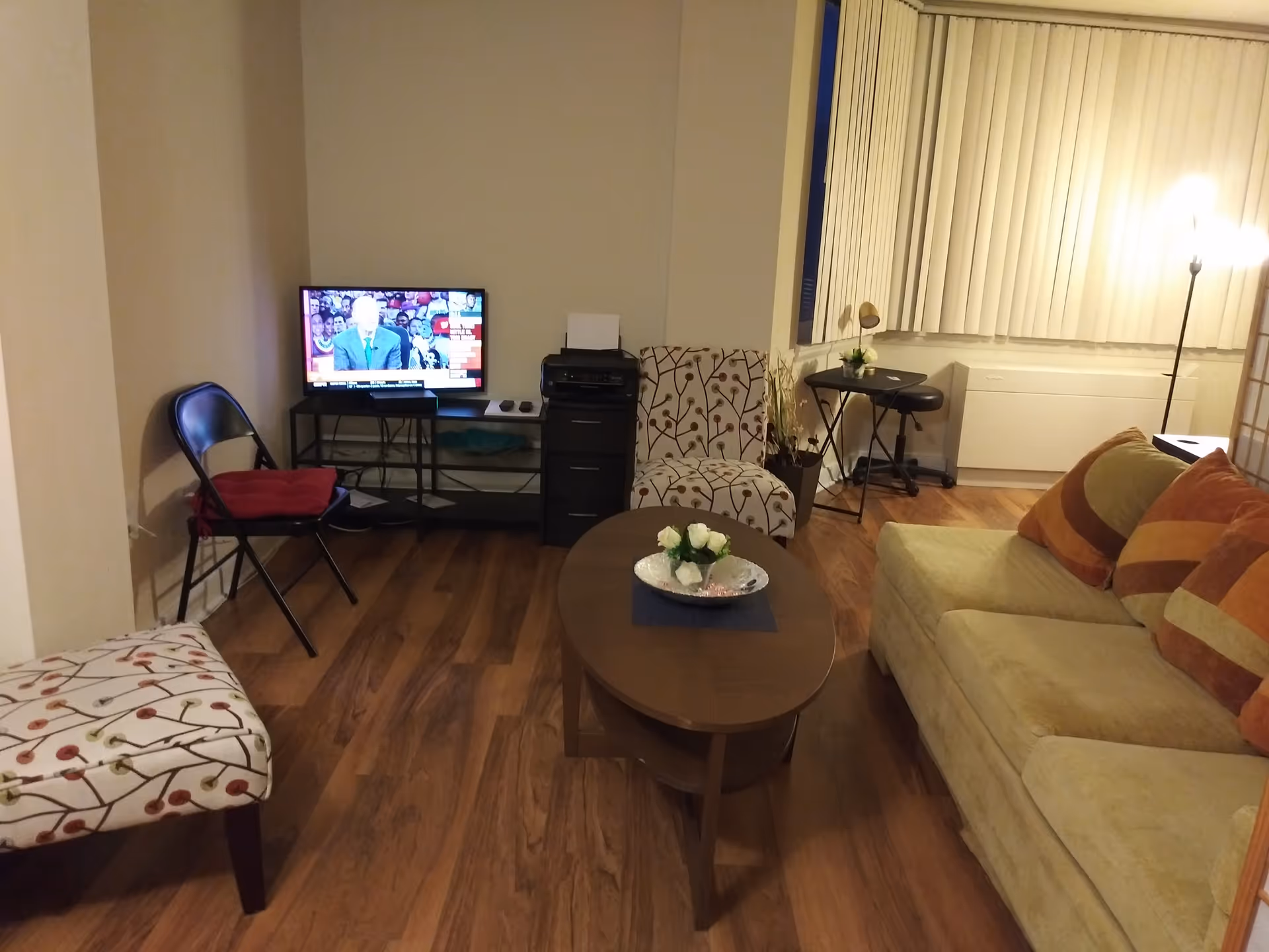 A cozy living room with a beige couch adorned with orange and brown striped pillows, a wooden coffee table with a small flower arrangement, two patterned armchairs, a black folding chair with a red cushion, a TV on a stand showing a news broadcast, a printer on a small black cabinet, a small table with a lamp and a stool near a window with vertical blinds, and a floor lamp providing warm lighting.